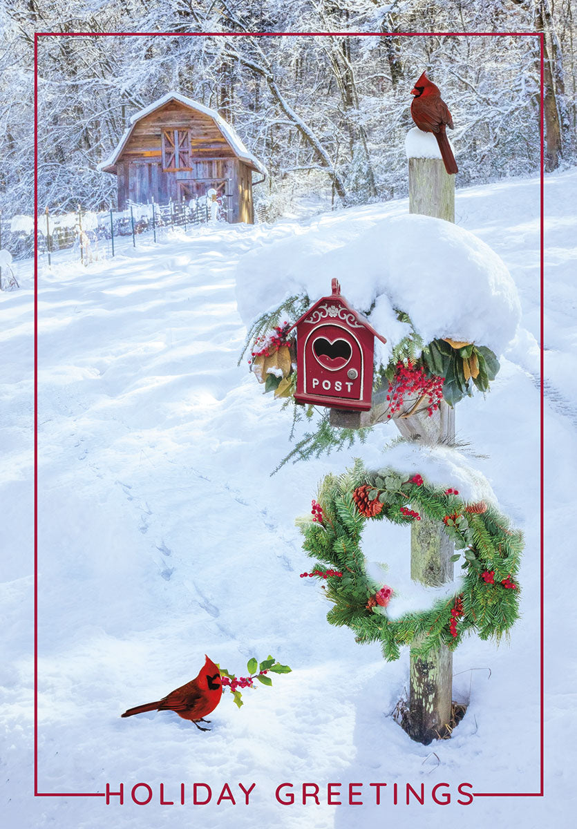 Front of a festive winter greeting card featuring a snowy rural landscape, two vibrant red cardinals, a rustic wooden barn, a charming red mailbox, and a green holiday wreath. Text reads 'HOLIDAY GREETINGS'.