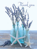 Close-up of a heartfelt thank you greeting card front, featuring dried lavender stems in four clear blue glass bottles, two white starfish, and a rustic light wood surface, with 'Thank you' in elegant script.