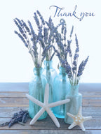 Close-up of a heartfelt thank you greeting card front, featuring dried lavender stems in four clear blue glass bottles, two white starfish, and a rustic light wood surface, with 'Thank you' in elegant script.