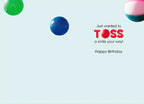 Inside view of a humorous birthday greeting card, showing an open light blue interior with floating colorful balls and the playful message 'Just wanted to TOSS a smile your way! Happy Birthday'.