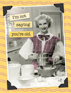 Funny greeting card front featuring a smiling retro woman sifting flour, with text 'I'm not saying you're old.' on a yellow background with wavy lines and black photo corners. Perfect for a humorous birthday message.