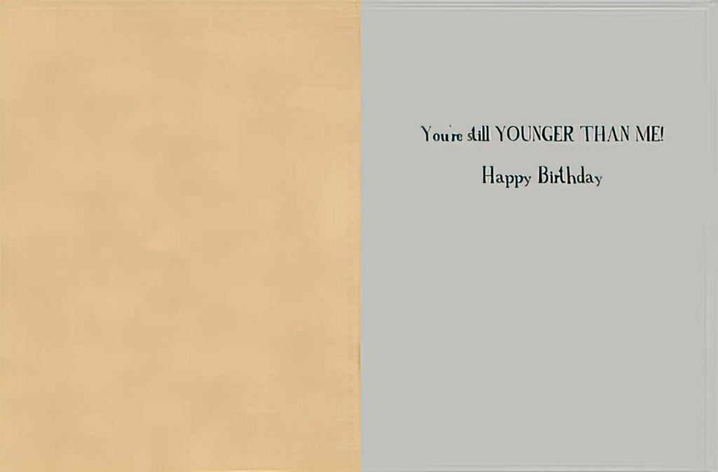Inside view of a humorous greeting card revealing the playful message 'You're still YOUNGER THAN ME! Happy Birthday' against a light gray and beige background, ideal for friends.