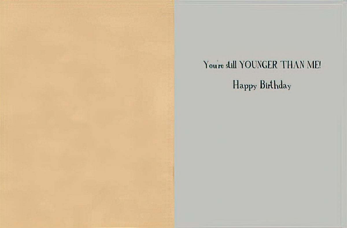 Inside view of a humorous greeting card revealing the playful message 'You're still YOUNGER THAN ME! Happy Birthday' against a light gray and beige background, ideal for friends.