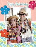 Front of a colorful anniversary greeting card featuring two children dressed as tourists with sunglasses, straw hats, and leis. They are smiling, holding a camera and a tropical drink, against a background of 'Happy Anniversary' text, blue, red, and orange flowers, and a blue and green swirl pattern, celebrating a spouse's special day.