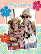 Front of a colorful anniversary greeting card featuring two children dressed as tourists with sunglasses, straw hats, and leis. They are smiling, holding a camera and a tropical drink, against a background of 'Happy Anniversary' text, blue, red, and orange flowers, and a blue and green swirl pattern, celebrating a spouse's special day.
