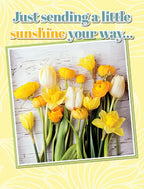 Bright yellow and white floral greeting card front with 'Just sending a little sunshine your way...' text, featuring a vibrant bouquet of tulips, daffodils, and ranunculus, perfect for encouraging a friend.