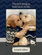 Front of a romantic anniversary greeting card featuring two golden retriever puppies snuggling under a patterned blanket. Text reads 'The best thing to hold on to in life... is each other.' on a dark blue chevron background, perfect for a spouse or partner.