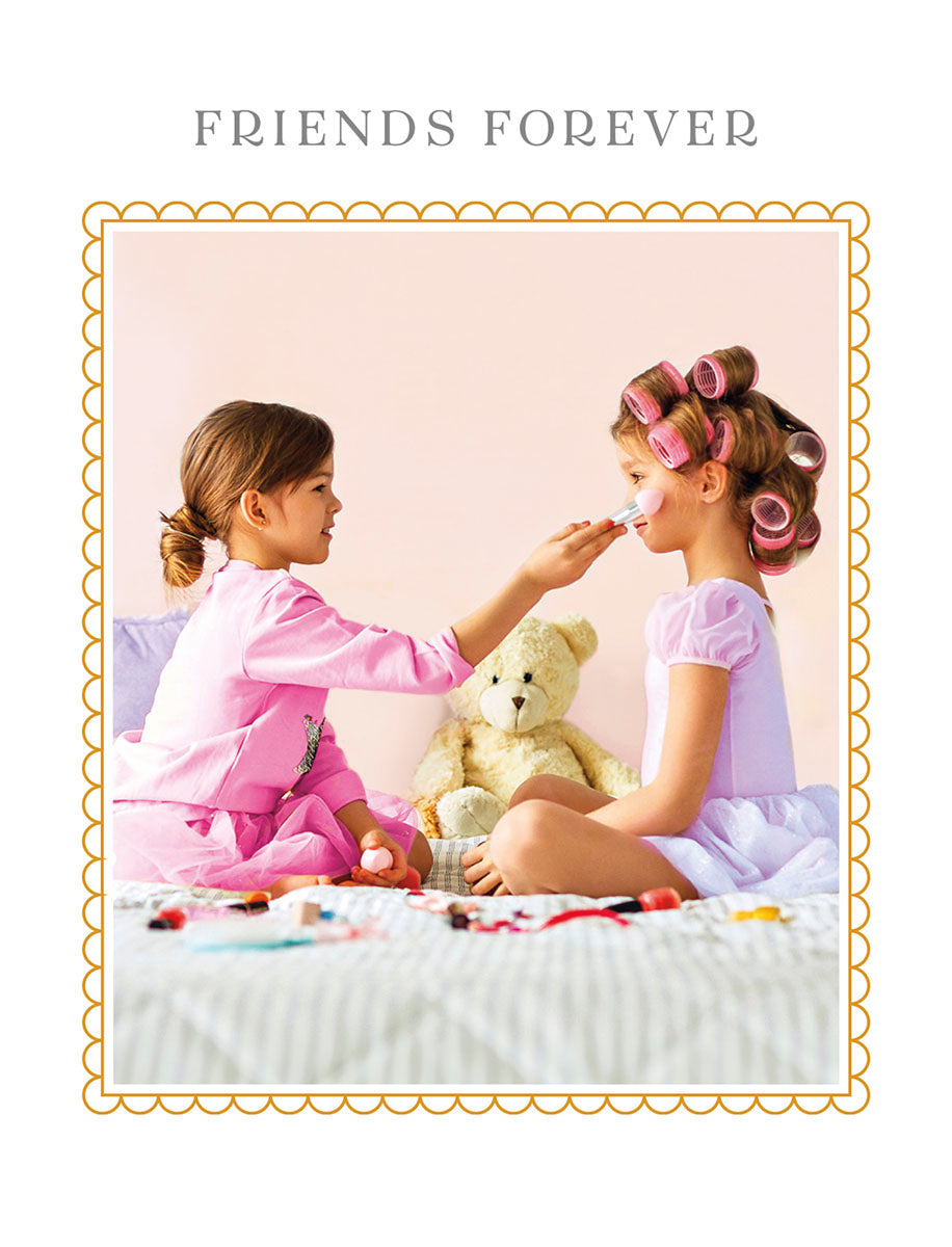 Adorable 'Friends Forever' greeting card featuring two happy young girls playing dress-up with makeup on a bed, alongside a teddy bear, framed by a whimsical border. Ideal for children's special moments.