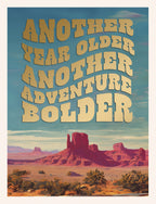 Front of an inspiring birthday card featuring a golden wavy text 'ANOTHER YEAR OLDER ANOTHER ADVENTURE BOLDER' against a stunning desert landscape with red rock formations and palm trees under a blue sky. Perfect for celebrating a friend's new chapter.