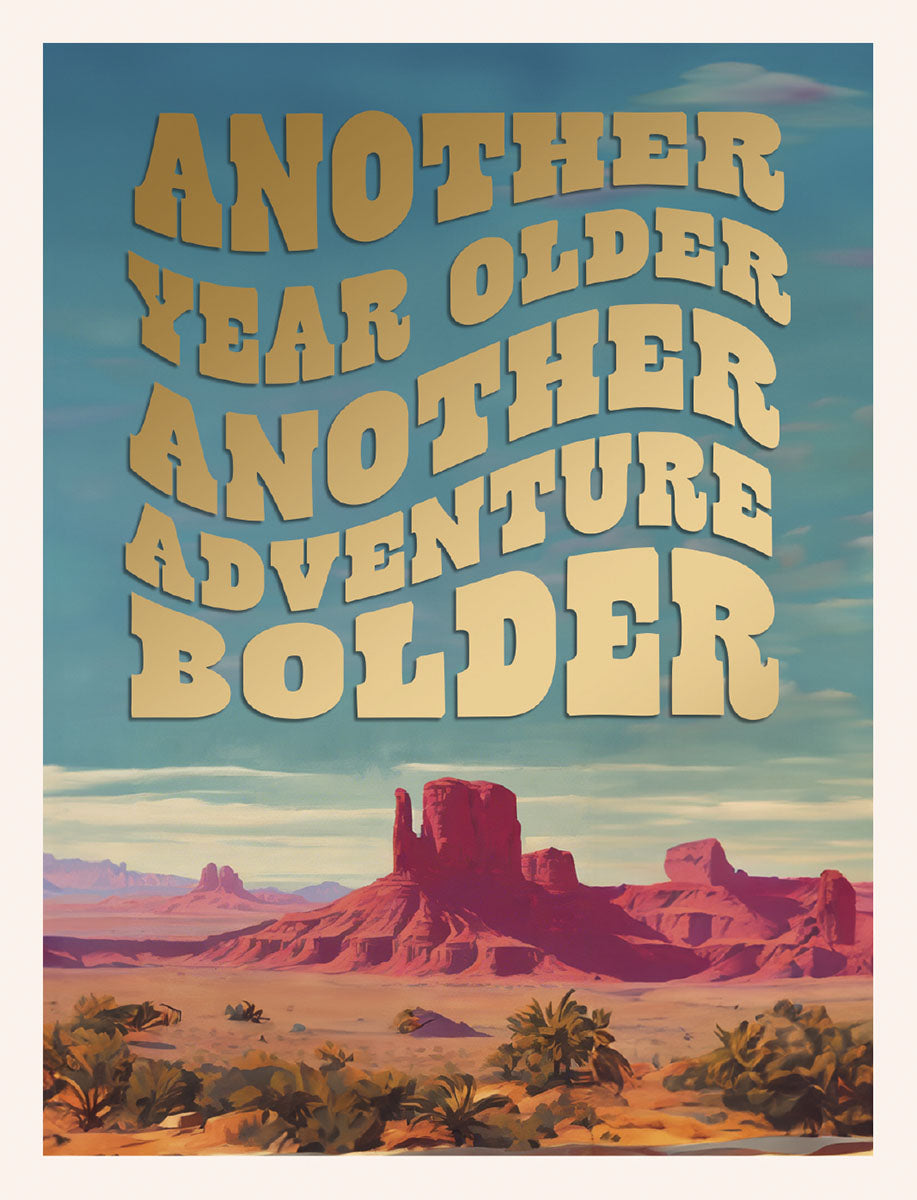 Front of an inspiring birthday card featuring a golden wavy text 'ANOTHER YEAR OLDER ANOTHER ADVENTURE BOLDER' against a stunning desert landscape with red rock formations and palm trees under a blue sky. Perfect for celebrating a friend's new chapter.
