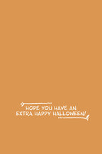 Inside of a cheerful fall greeting card with a warm orange background, featuring the white script message 'Hope you have an extra happy Halloween!' and decorative flourishes.
