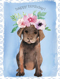 Front of a watercolor birthday greeting card featuring a charming brown puppy wearing a vibrant pink and purple floral crown, set against a soft blue background with delicate patterned borders and 'happy birthday!' text.