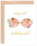Front of a white birthday greeting card featuring a watercolor illustration of pink-lensed round sunglasses adorned with soft pink, blue, and green floral accents. Above the sunglasses, the text reads "stay cool" and below, "birthday girl!" in elegant gold script. A brown kraft paper envelope is visible behind the card.