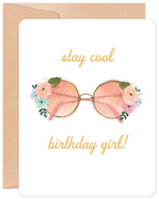 Front of a white birthday greeting card featuring a watercolor illustration of pink-lensed round sunglasses adorned with soft pink, blue, and green floral accents. Above the sunglasses, the text reads "stay cool" and below, "birthday girl!" in elegant gold script. A brown kraft paper envelope is visible behind the card.