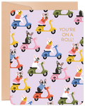 Front of a cute birthday card featuring multiple French Bulldogs in various colored outfits riding vibrant scooters on a lavender background. The text 'YOU'RE ON A ROLL!' is prominently displayed in orange. A kraft paper border is visible.