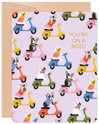 Front of a cute birthday card featuring multiple French Bulldogs in various colored outfits riding vibrant scooters on a lavender background. The text 'YOU'RE ON A ROLL!' is prominently displayed in orange. A kraft paper border is visible.