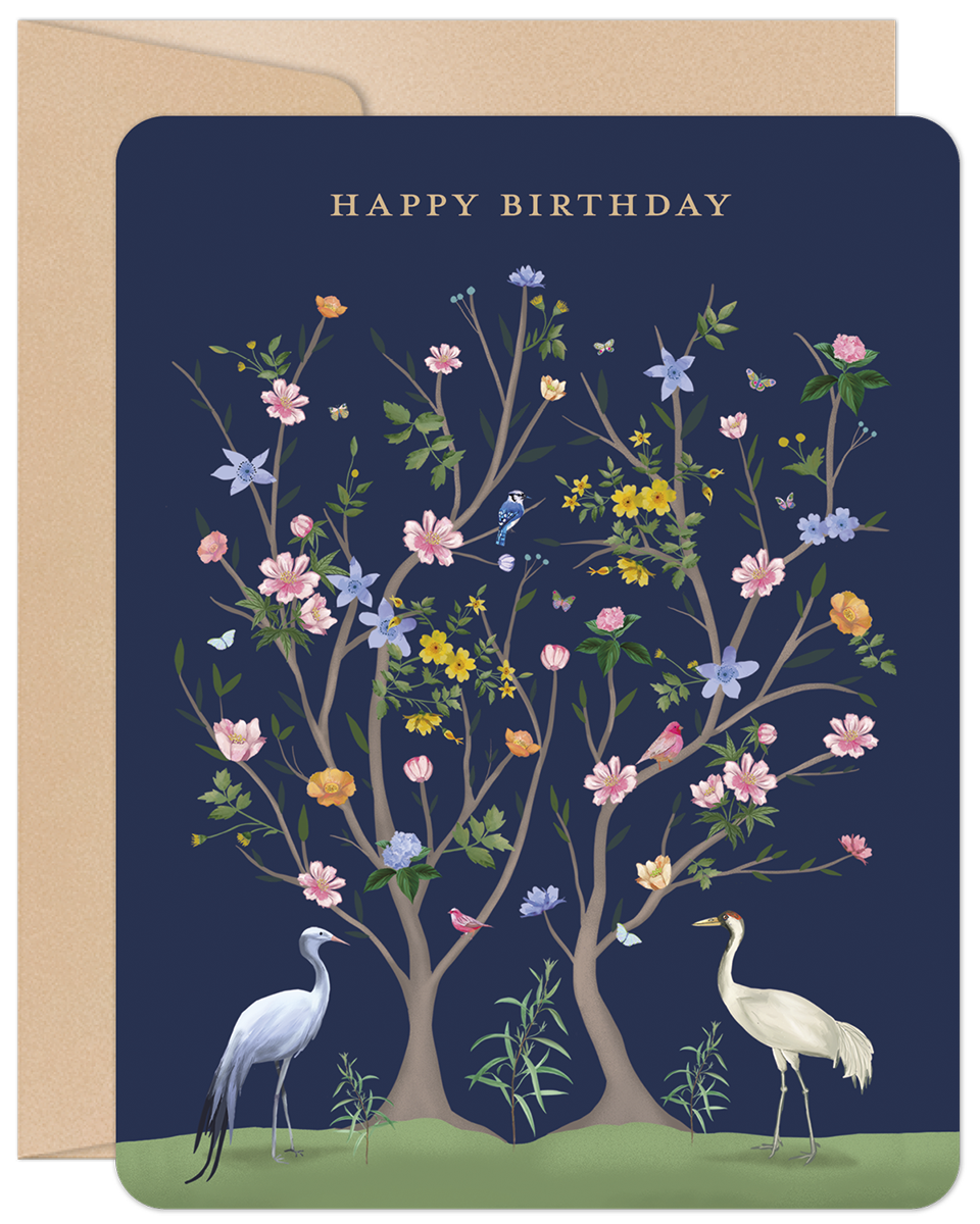 Elegant dark blue birthday greeting card featuring a detailed illustration of two white cranes and flourishing trees with various pink, yellow, and blue flowers, green leaves, and small birds, with 'HAPPY BIRTHDAY' in gold text, accompanied by a natural kraft envelope.