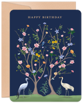 Elegant dark blue birthday greeting card featuring a detailed illustration of two white cranes and flourishing trees with various pink, yellow, and blue flowers, green leaves, and small birds, with 'HAPPY BIRTHDAY' in gold text, accompanied by a natural kraft envelope.