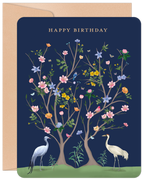 Elegant dark blue birthday greeting card featuring a detailed illustration of two white cranes and flourishing trees with various pink, yellow, and blue flowers, green leaves, and small birds, with 'HAPPY BIRTHDAY' in gold text, accompanied by a natural kraft envelope.
