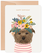 Cute brown terrier dog wearing a vibrant floral crown with yellow, pink, and green flowers, on a cream greeting card with 'HAPPY BIRTHDAY' text, perfect for a friend.