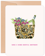 Watercolor illustration of a leopard print tote bag overflowing with vibrant purple and pink flowers, greenery, and a bottle of sparkling wine. The text 'HAVE A WINE-DERFUL BIRTHDAY' is printed below, set against a light background with a kraft brown envelope peeking out.