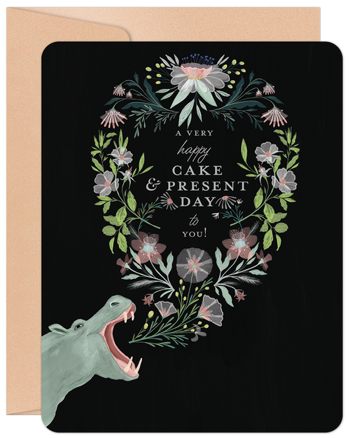 Elegant black greeting card featuring a charming hippo illustration with its mouth open, surrounded by a delicate oval wreath of pink, white, and green flowers and leaves. The text 'A very happy cake & present day to you!' is displayed within the floral frame, emphasizing a lighthearted celebration message. A kraft paper envelope peeks out from behind the card, suggesting a complete gift.
