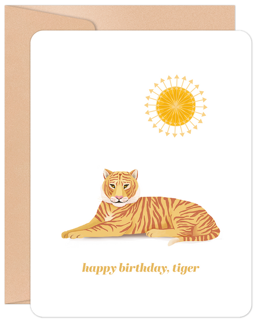 Front of a white greeting card featuring a stylized orange tiger laying down, with a golden sun icon above it. The text 'happy birthday, tiger' is printed below the tiger. A kraft brown envelope is visible behind the card.