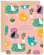 Front of a cute birthday greeting card featuring an array of white, black, and ginger cats lounging on colorful floral and patterned armchairs and sofas, surrounded by potted plants on a light pink background. The text 'HAPPY BIRTHDAY' is centrally placed.
