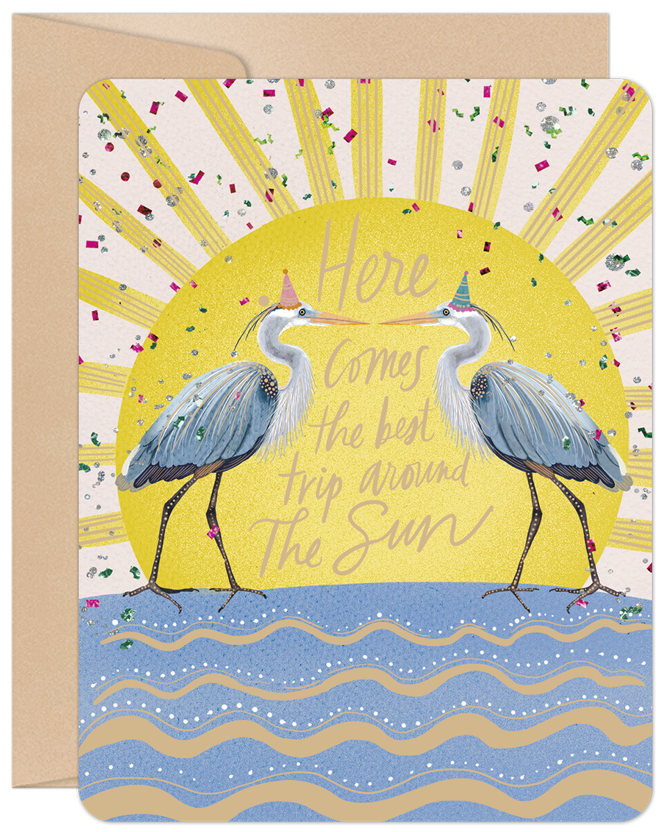 Vibrant greeting card featuring two elegant blue herons wearing festive party hats, standing on whimsical blue waves against a brilliant yellow sun with radiating golden rays and sparkling confetti. The card's front sentiment reads: 'Here comes the best trip around The Sun'.