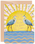 Whimsical birthday greeting card featuring two blue herons wearing festive party hats, set against a shimmering yellow sunburst with colorful confetti, above blue and white ocean waves. The card front reads 'Here Comes the best trip around The Sun' and is accompanied by a kraft paper envelope.
