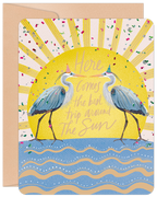 Whimsical birthday greeting card featuring two blue herons wearing festive party hats, set against a shimmering yellow sunburst with colorful confetti, above blue and white ocean waves. The card front reads 'Here Comes the best trip around The Sun' and is accompanied by a kraft paper envelope.