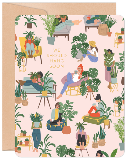 Charming friendship card front with diverse women relaxing among houseplants on pink. Text: 'We Should Hang Soon'. Perfect for plant lovers.