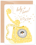 Front view of a cute new baby greeting card featuring a yellow vintage telephone, hearts, and text 'baby is here! xoxo' on a white background, accompanied by a brown envelope.