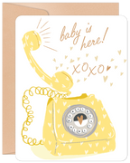 Front view of a cute new baby greeting card featuring a yellow vintage telephone, hearts, and text 'baby is here! xoxo' on a white background, accompanied by a brown envelope.