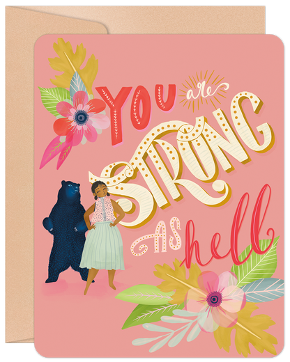 Front of a pink greeting card featuring a woman and a bear with colorful floral accents. Text: "YOU are STRONG AS hell".