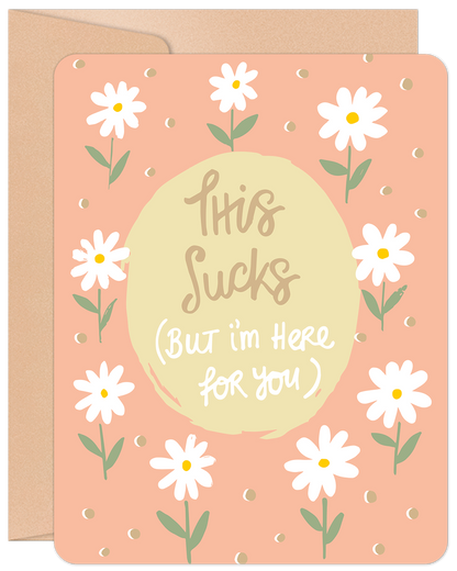 Greeting card with white daisies and text 'This Sucks (But I'm Here for You)' on a peach background, with a kraft envelope.