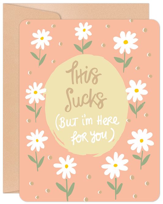 Greeting card with white daisies and text 'This Sucks (But I'm Here for You)' on a peach background, with a kraft envelope.
