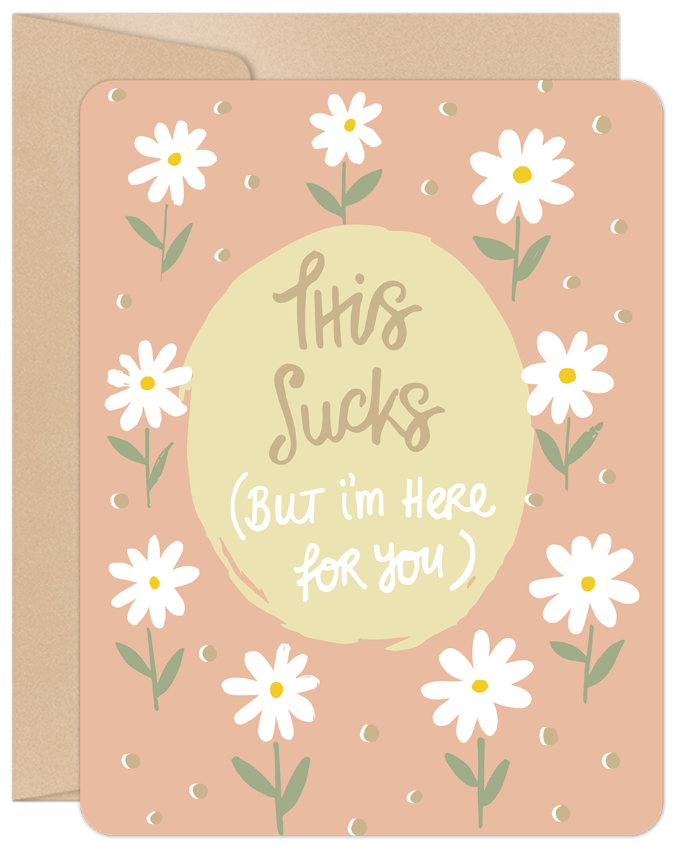 Front view of an encouragement greeting card featuring white daisies on a peach background with the handwritten text 'This Sucks (But I'm Here for You)' in a central light yellow circle, ideal for offering support.