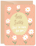 Front view of an encouragement greeting card featuring white daisies on a peach background with the handwritten text 'This Sucks (But I'm Here for You)' in a central light yellow circle, ideal for offering support.