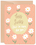 Front view of an encouragement greeting card featuring white daisies on a peach background with the handwritten text 'This Sucks (But I'm Here for You)' in a central light yellow circle, ideal for offering support.