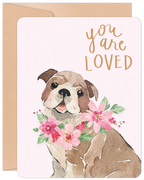 Front of a greeting card featuring a watercolor illustration of a cute bulldog wearing a pink floral wreath. Gold script above reads 'you are LOVED'. The card has rounded corners and a slight pink tint, with a kraft envelope visible behind it.