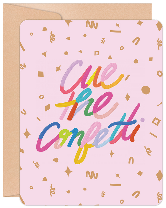 Front of a pink greeting card with colorful 'Cue the Confetti' text and gold confetti shapes, accompanied by a kraft envelope.