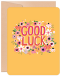 Vibrant yellow greeting card with 'GOOD LUCK' in pink letters, surrounded by a cheerful wreath of colorful flowers, with a brown envelope peeking behind.