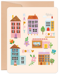 Front of a cheerful new home greeting card featuring an illustration of colorful houses, trees, and flowers with the words 'HAPPY COZY HOME' on a light beige background.