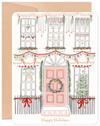 Close-up of a festive holiday greeting card featuring an illustrated white house with a pink door, adorned with green garlands, red bows, string lights, and wreaths, all against a kraft paper envelope.