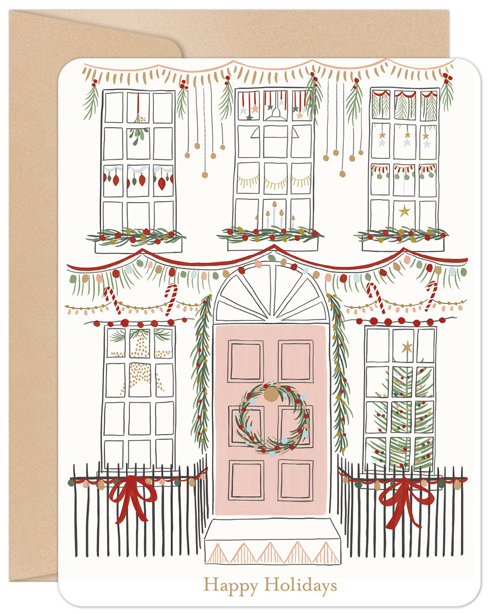 Close-up of a festive holiday greeting card featuring an illustrated white house with a pink door, adorned with green garlands, red bows, string lights, and wreaths, all against a kraft paper envelope.