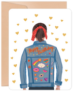Cute birthday greeting card for her, featuring a person from behind wearing a blue denim jacket adorned with colorful patches like a rainbow, donut, watermelon, and stars, with 'Happy Birthday' written across the back, against a white background with gold hearts, alongside a craft paper envelope.