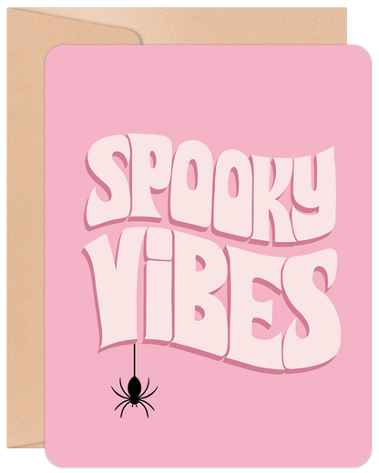 Pink Halloween greeting card featuring "Spooky Vibes" text in white and a black spider, resting on a brown kraft envelope.