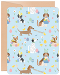 Front of an Easter greeting card featuring a light blue background patterned with various cute cartoon dogs in bunny ears, colorful decorated eggs, and yellow chicks. Dog breeds include Beagle, Boston Terrier, Dachshund, Dalmatian, and Corgi. Text reads "HAPPY EASTER".