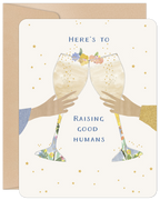 Front view of a cream-colored greeting card featuring two hands holding champagne flutes in a celebratory toast, adorned with delicate floral details and scattered gold stars. The text reads "HERE'S TO RAISING GOOD HUMANS".